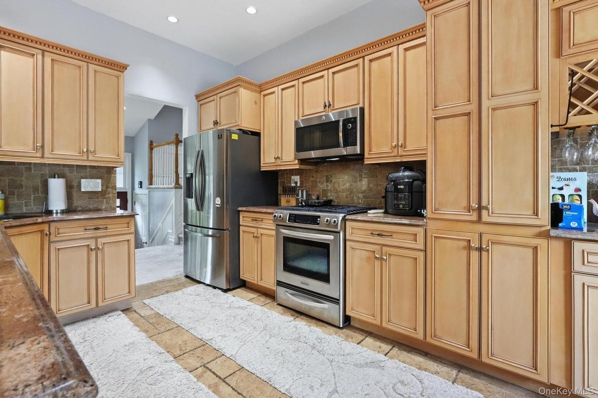 Kitchen featuring light wood cabinetry with crown molding, stainless steel appliances, stone tile flooring, and a tiled backsplash