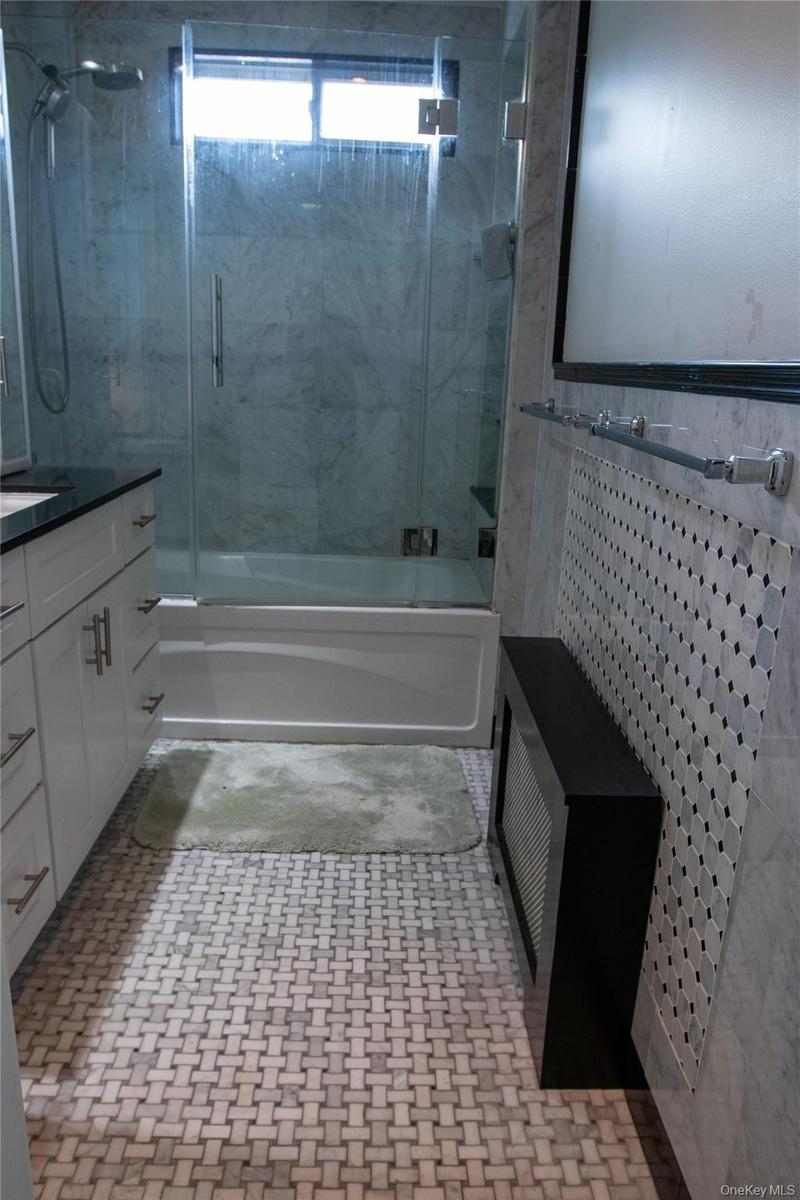 Bathroom featuring a glass-enclosed shower over a tub, white vanity cabinetry with dark countertops, a mosaic tile accent wall, and woven pattern floo