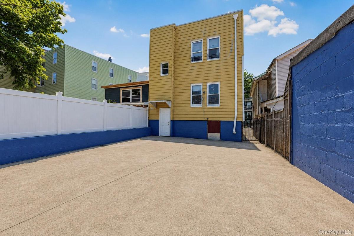 Paved outdoor space featuring a yellow-sided structure with multiple windows, a white privacy fence, and a blue painted foundation