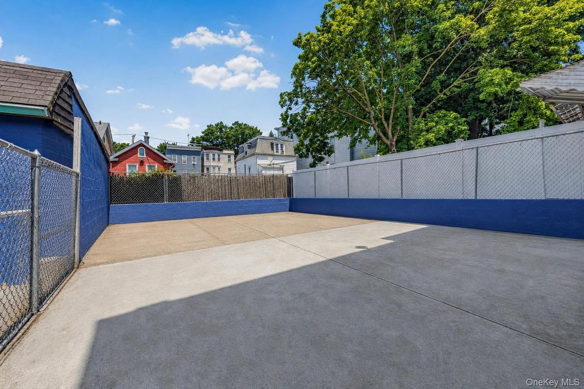 Paved outdoor space featuring a yellow-sided structure with multiple windows, a white privacy fence, and a blue painted foundation