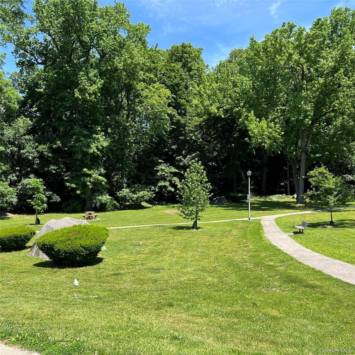 Expansive green space featuring a paved walking path, mature trees, manicured hedges, and a picnic tables