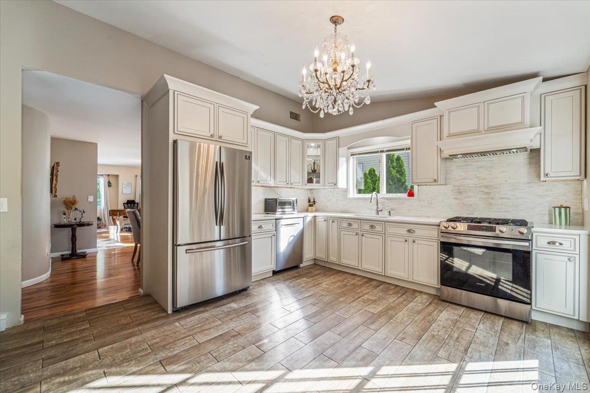 Kitchen featuring white cabinetry, stainless steel appliances, a crystal chandelier, wood-finish flooring, and a white tile backsplash