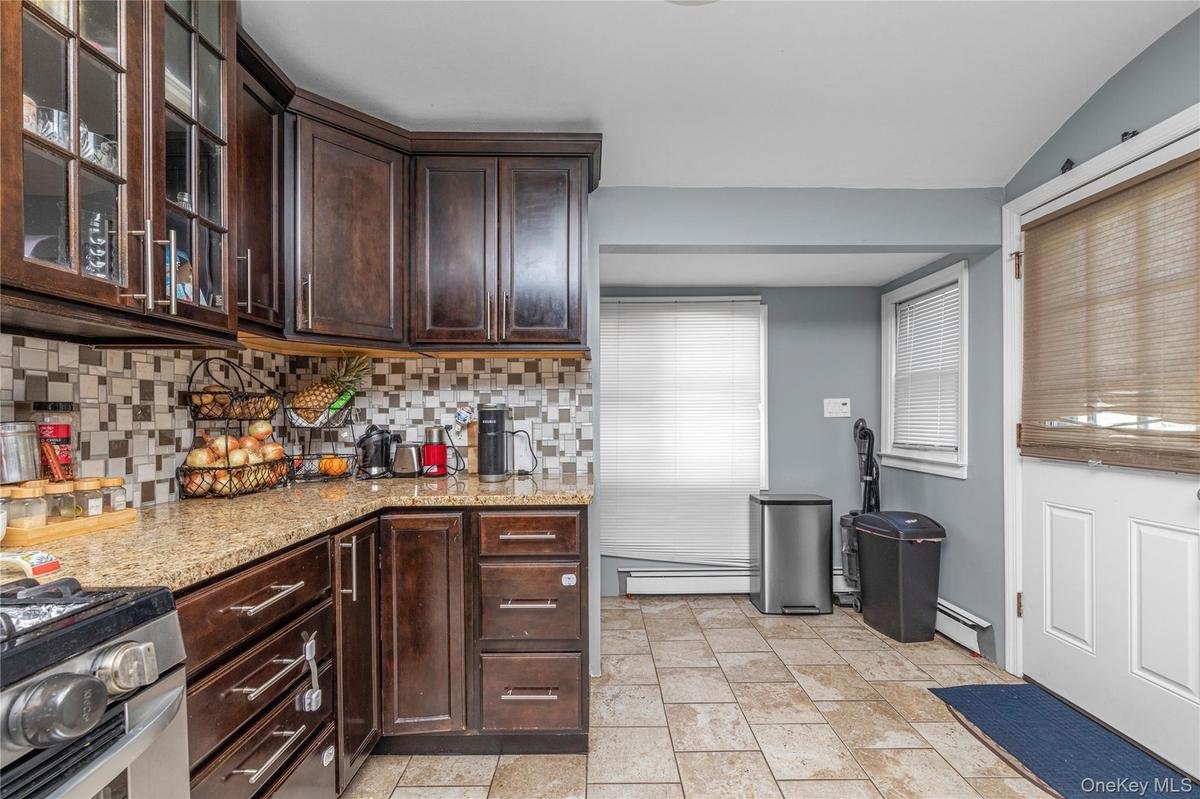 Kitchen featuring dark brown cabinetry, light stone counters, decorative backsplash, a baseboard radiator, and glass insert cabinets
