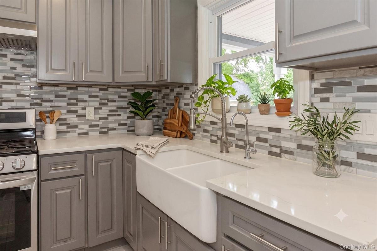 Modern kitchen featuring a white farmhouse-style sink, light gray cabinetry, stainless steel appliances, a mosaic tile backsplash, and light-colored c
