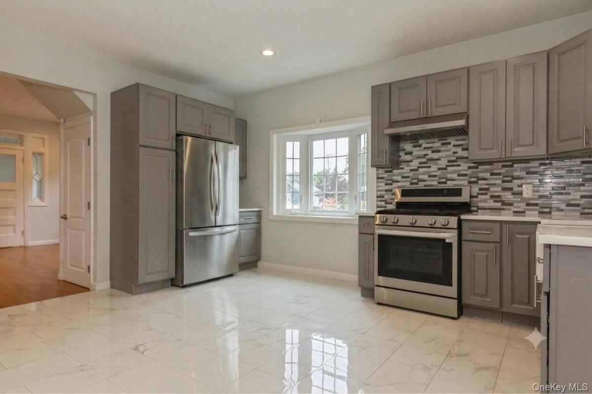 Kitchen featuring white marble-finish tile flooring, grey cabinetry, stainless steel appliances, a multi-tone tile backsplash, and a bay window