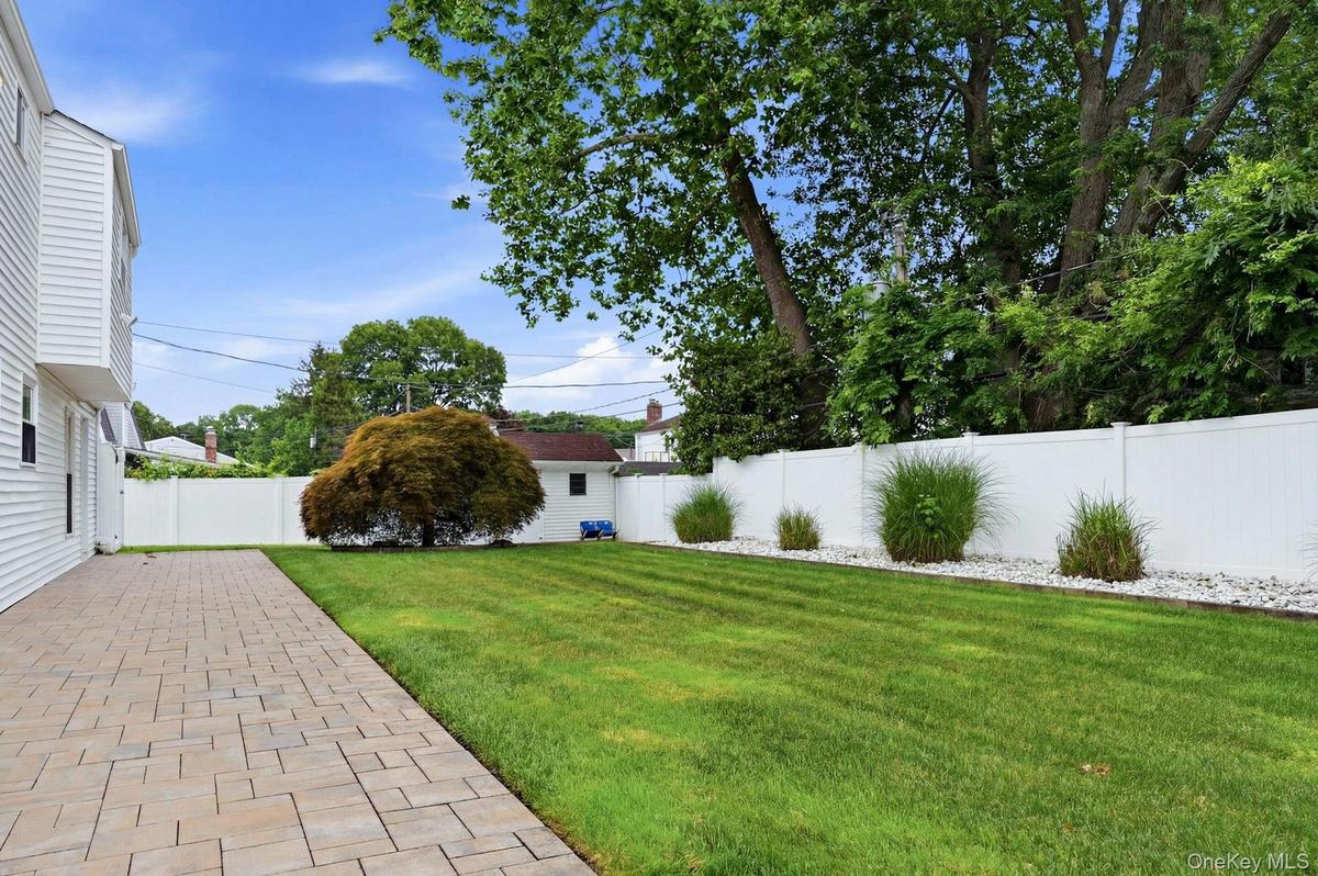 Expansive backyard featuring a paver patio, manicured lawn, white privacy fence, and mature landscaping