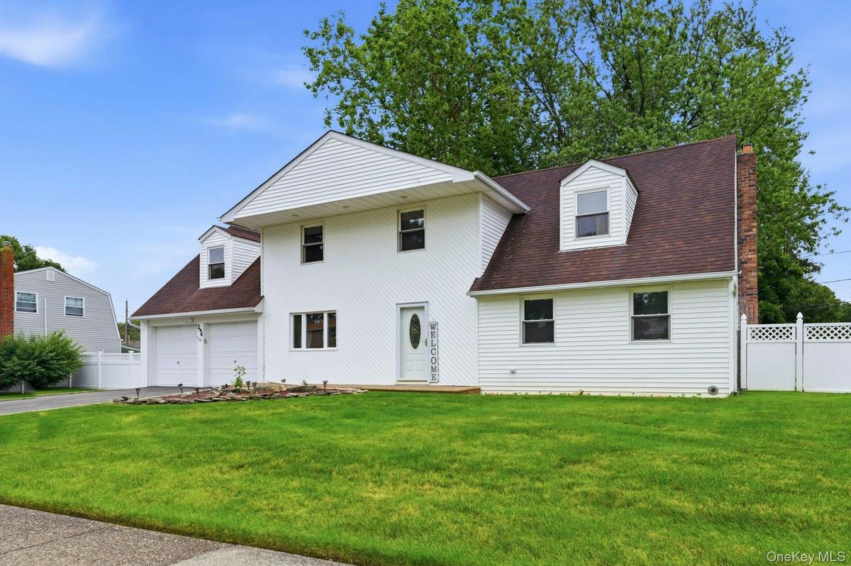 White siding exterior with a brown shingle roof, featuring a two-car garage, dormer window, front porch, and a white lattice fence