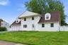 White siding exterior with a brown shingle roof, featuring a two-car garage, dormer window, front porch, and a white lattice fence