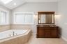 Soaking jet tub with tiled surround, dual-sink vanity with stone countertop, decorative windows, and vaulted ceiling with recessed lighting and skylig