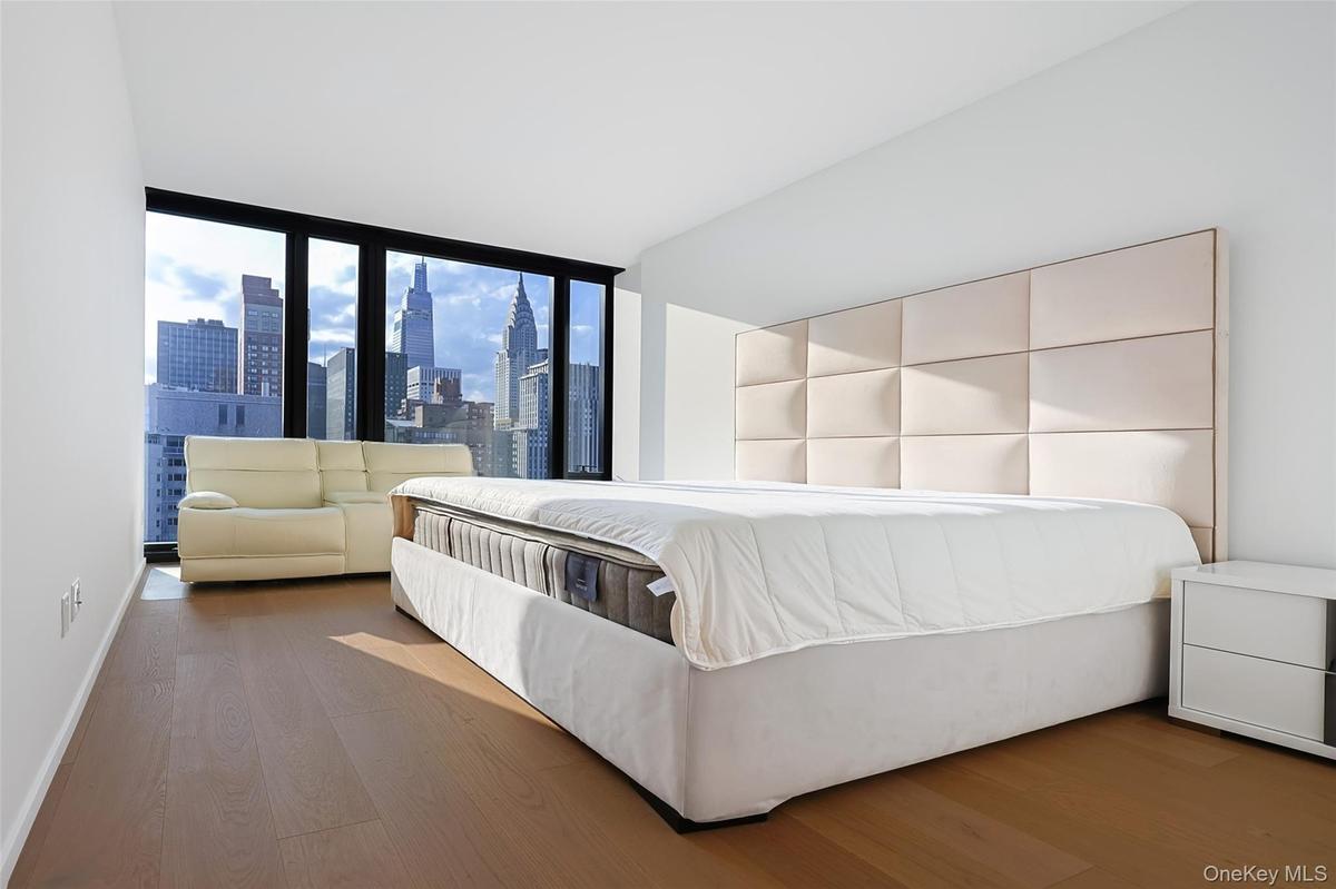 Primary Bedroom with Expansive windows showcasing cityscape views, light wood-finish flooring, white walls, and a paneled upholstered headboard