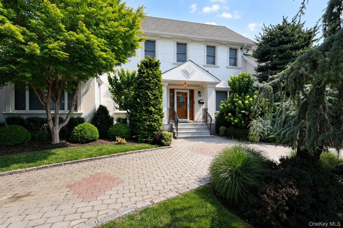 White stucco exterior with a gray shingle roof, featuring a paver driveway, mature landscaping, a covered entryway, and a natural wood front door