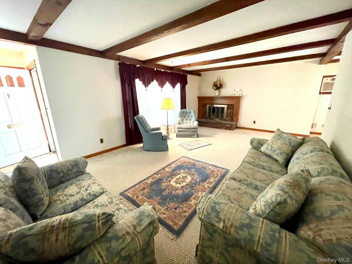 Living area featuring exposed wood beams, a prominent fireplace with a wood mantle and stone hearth, and light-colored carpeting