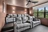 Dedicated media room featuring tiered seating, built-in wall sconces, a modern ceiling fan, and large black-framed windows overlooking mature trees