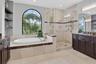 Spacious bathroom featuring an arched window, a built-in soaking tub, a glass-enclosed shower, dark wood cabinetry, and stone countertops