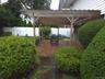 Brick paver patio featuring a wooden pergola with translucent roofing panels, surrounded by a manicured lawn, mature shrubs, and fencing