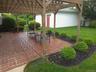 Brick paver patio featuring a wooden pergola with translucent roofing panels, surrounded by a manicured lawn, mature shrubs, and fencing