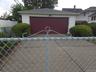 Detached and gated 2 car garage featuring white horizontal siding, a dark red garage door, dark red side door and gabled roof