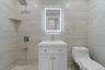 Bathroom featuring large format light-toned tile, a rectangular illuminated mirror, a white vanity with under-sink storage, a wall-mounted shower syst