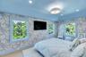 Spacious bedroom featuring light wood-finish flooring, patterned wallpaper, a built-in wall-mounted television, and multiple windows with light blue t