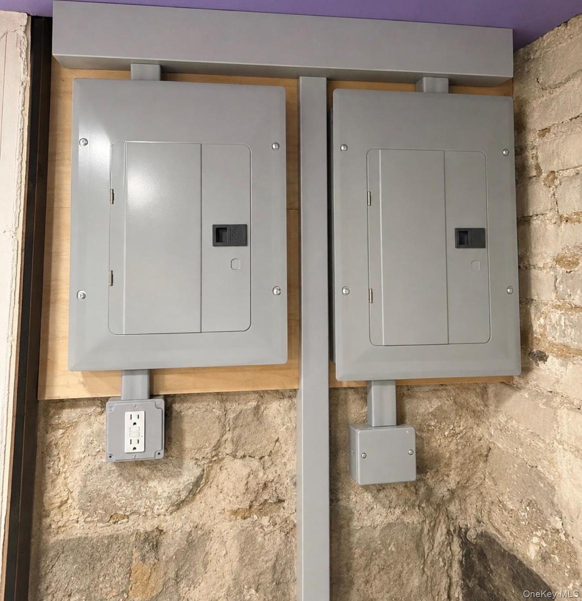 Electrical panels with a wall outlet, mounted on a wood-finish backboard, set against a natural stone wall