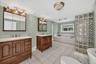 Dual wood-finish vanities with integrated sinks, a built-in soaking tub, tile flooring, and a glass block privacy wall