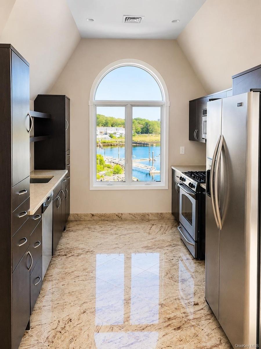 Arched window with water scenery, dark wood-finish cabinetry, stainless steel appliances, light-toned countertop, and polished tile flooring