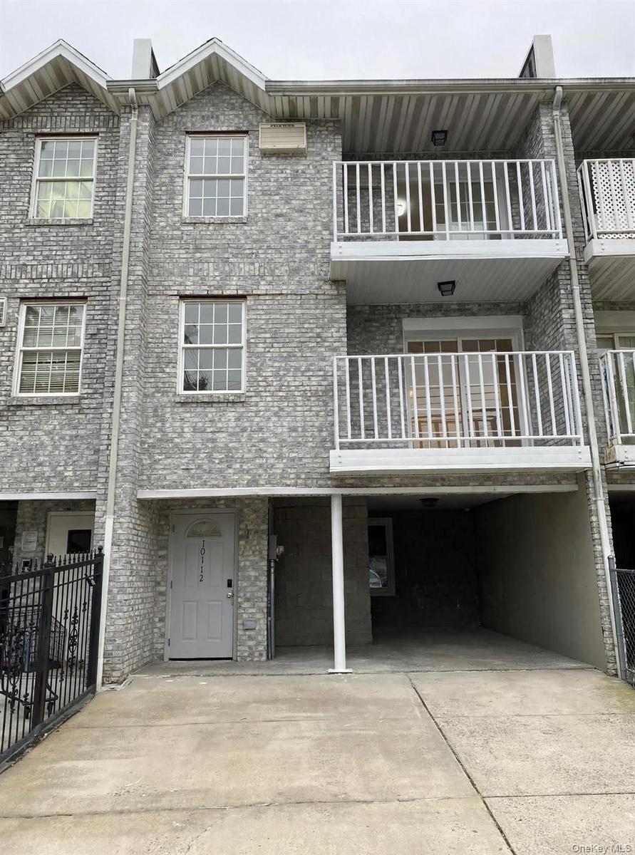 Multi-level brick facade featuring two stacked balconies with white metal railings, a covered parking area, and multi-pane windows