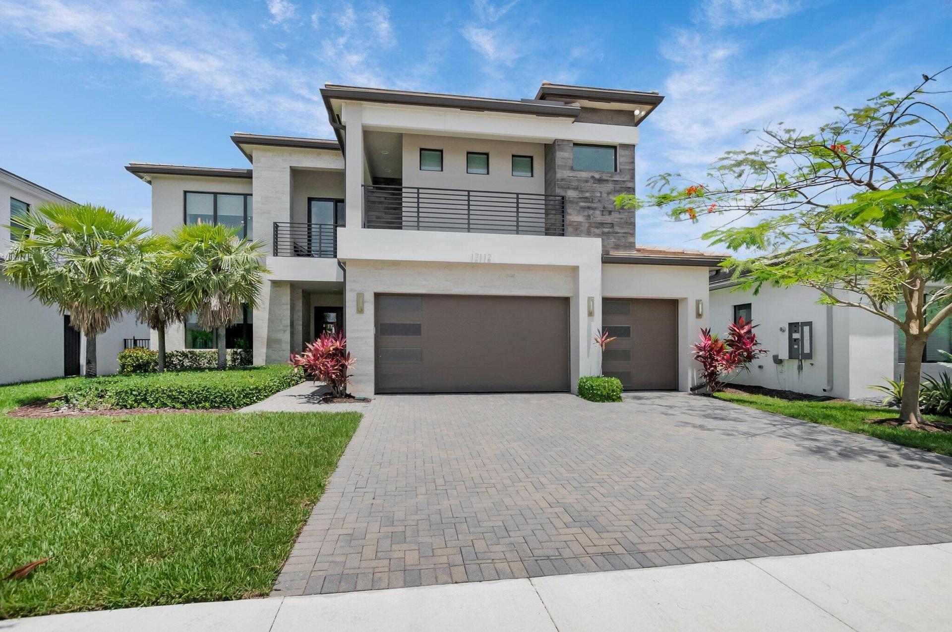 Contemporary two-story residence featuring a paver driveway, multiple garage doors, a second-story balcony with metal railing, and a stone accent faca