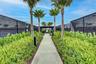 Paved walkway flanked by lush green landscaping and mature palm trees, leading to covered seating areas and tennis courts with black chain-link fencin