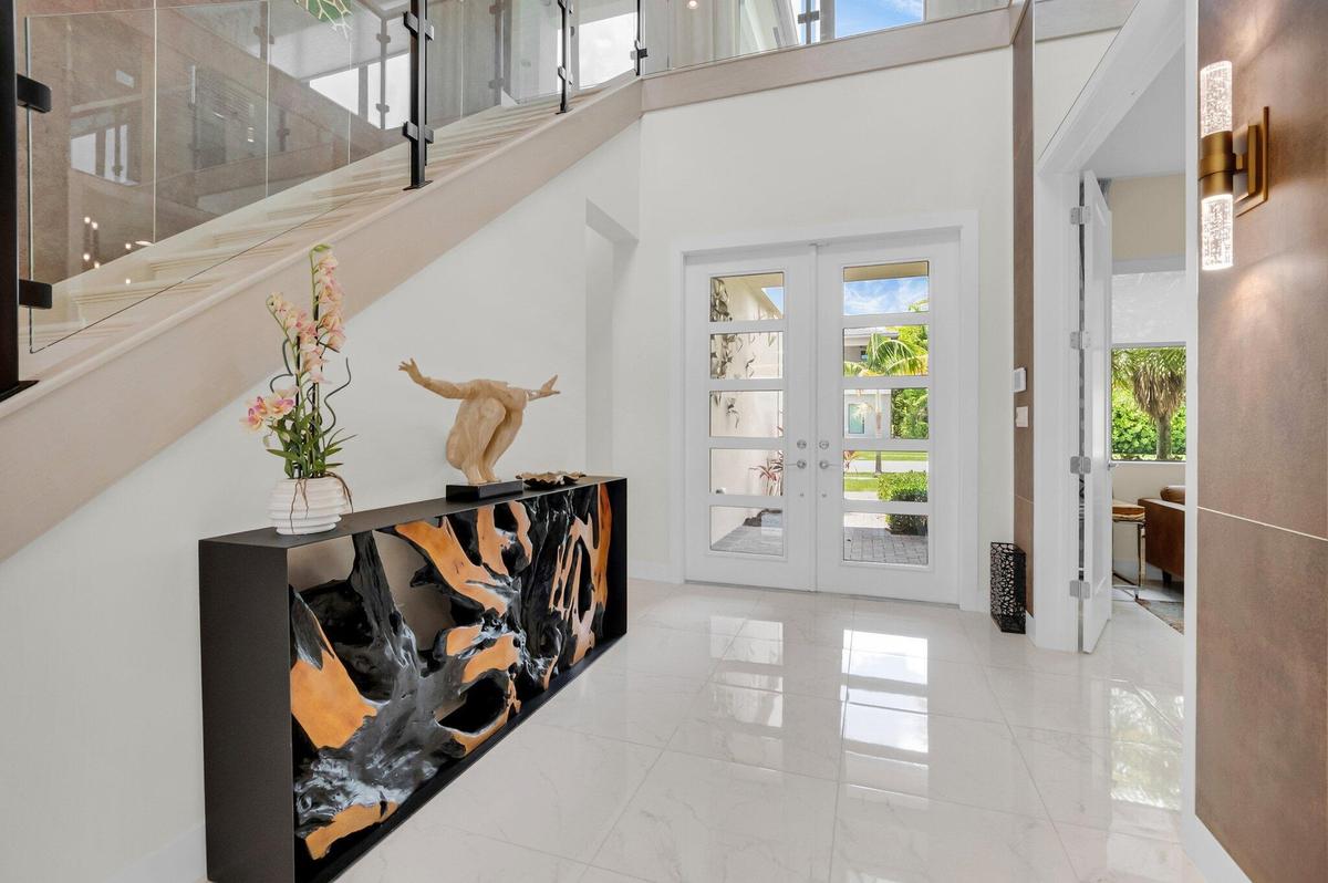 Double entry doors with multiple glass panels, a contemporary console table, polished tile flooring, a staircase with glass railings, and a wall-mount