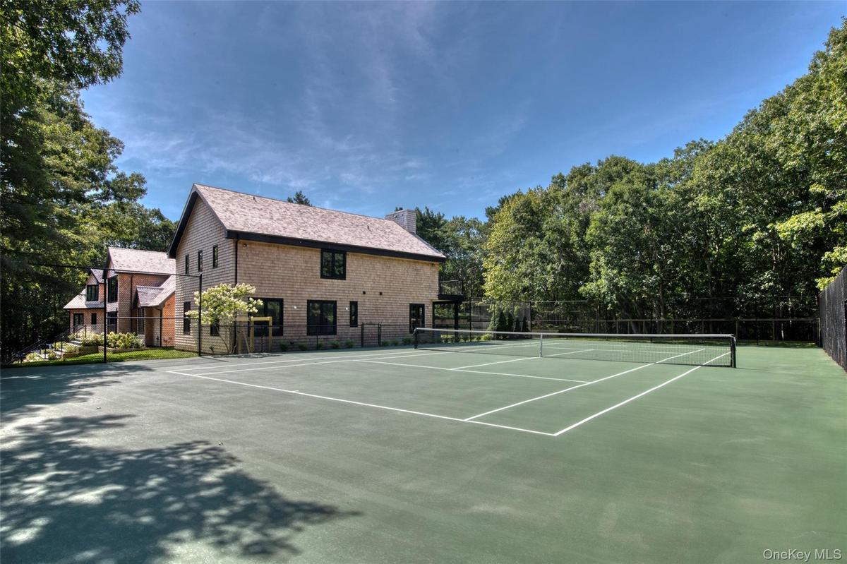 Full-size tennis court with a perimeter fence, adjacent to a residence with shingle siding and dark-trimmed windows