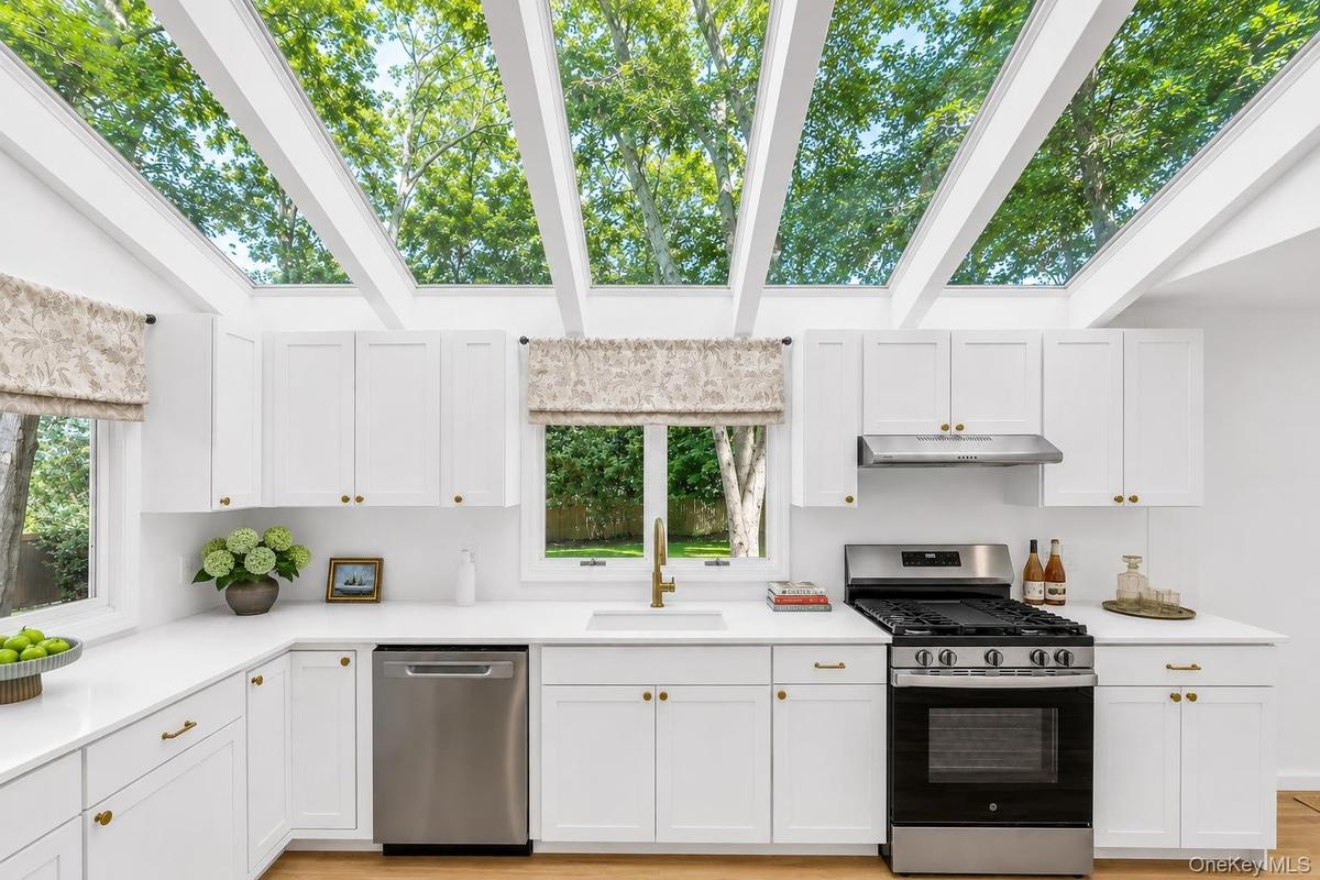 White cabinetry with brass hardware, stainless steel appliances, light wood-finish flooring, and a vaulted ceiling with exposed beams and skylights