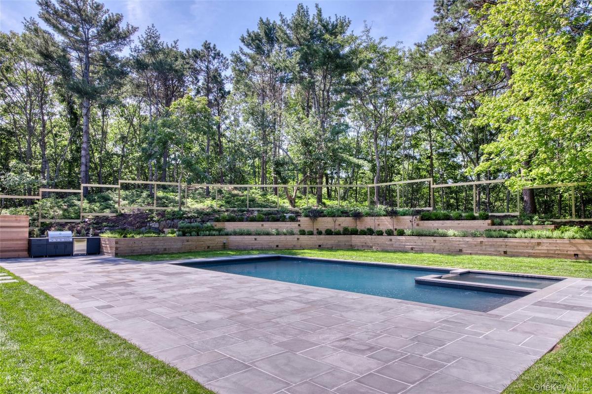 Swimming pool with integrated spa, expansive paver patio, built-in outdoor kitchen, tiered landscaping, and mature perimeter trees