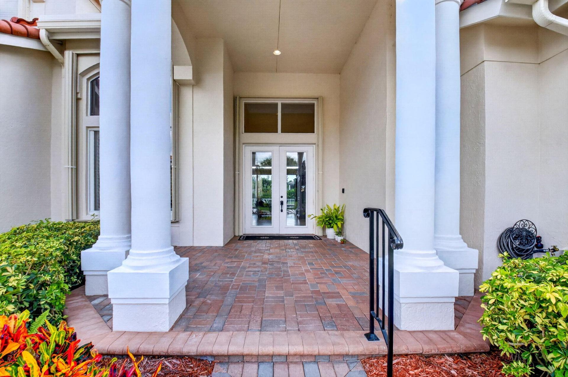 Portico entry featuring large columns, double glass-paneled doors, brick paver flooring, and a wrought iron handrail