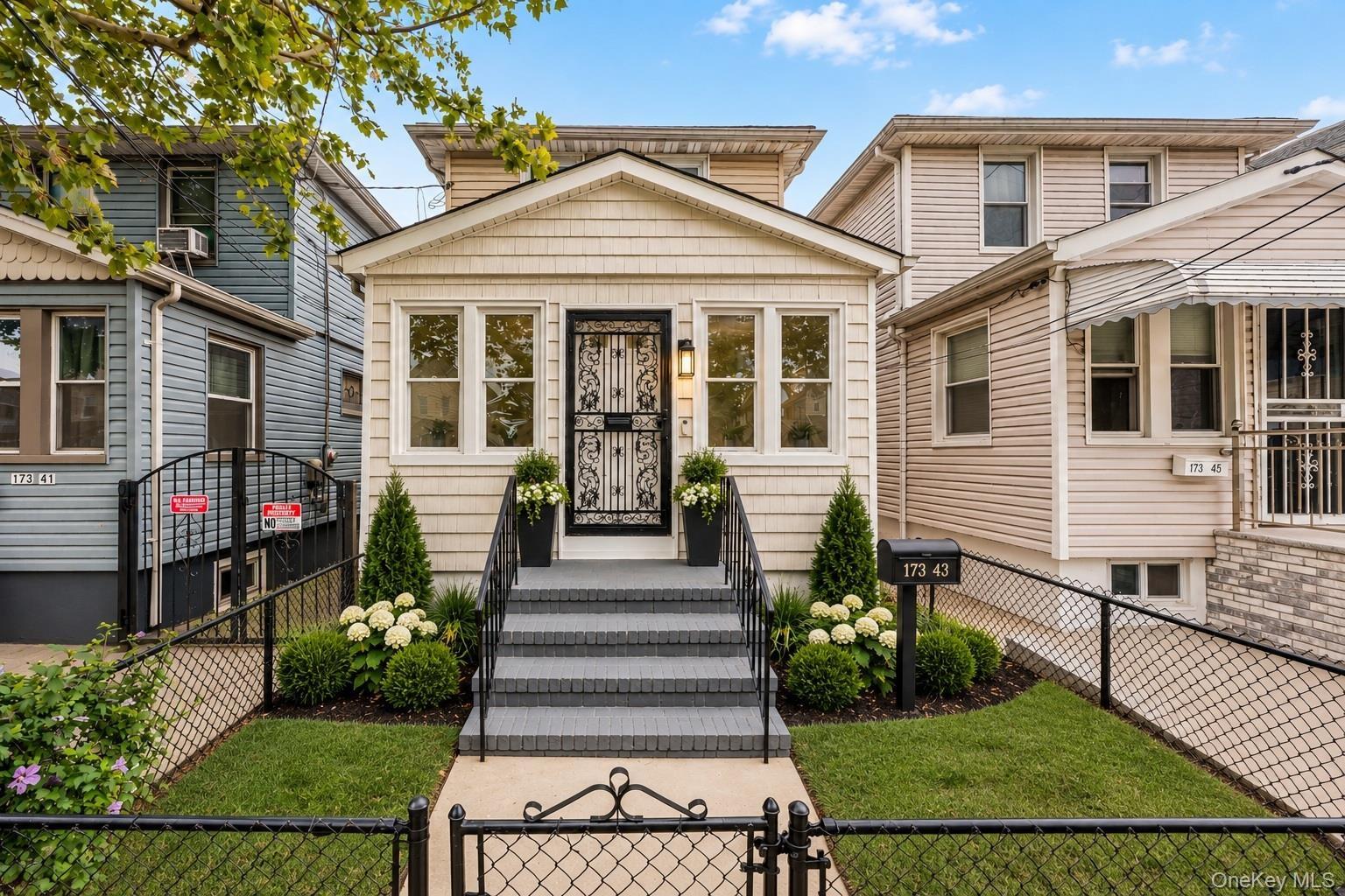 Paved entryway leading to a staircase with black railings, a black security door, and a manicured front yard with mature plantings and a wrought iron 