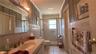 Vanity with integrated sink, brass fixtures, mirrored medicine cabinet, tiled shower with glass enclosure, and a window