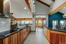 Vaulted ceilings with exposed wood beams, wood-finish cabinetry, dark countertops, a stainless steel range hood, and tile flooring