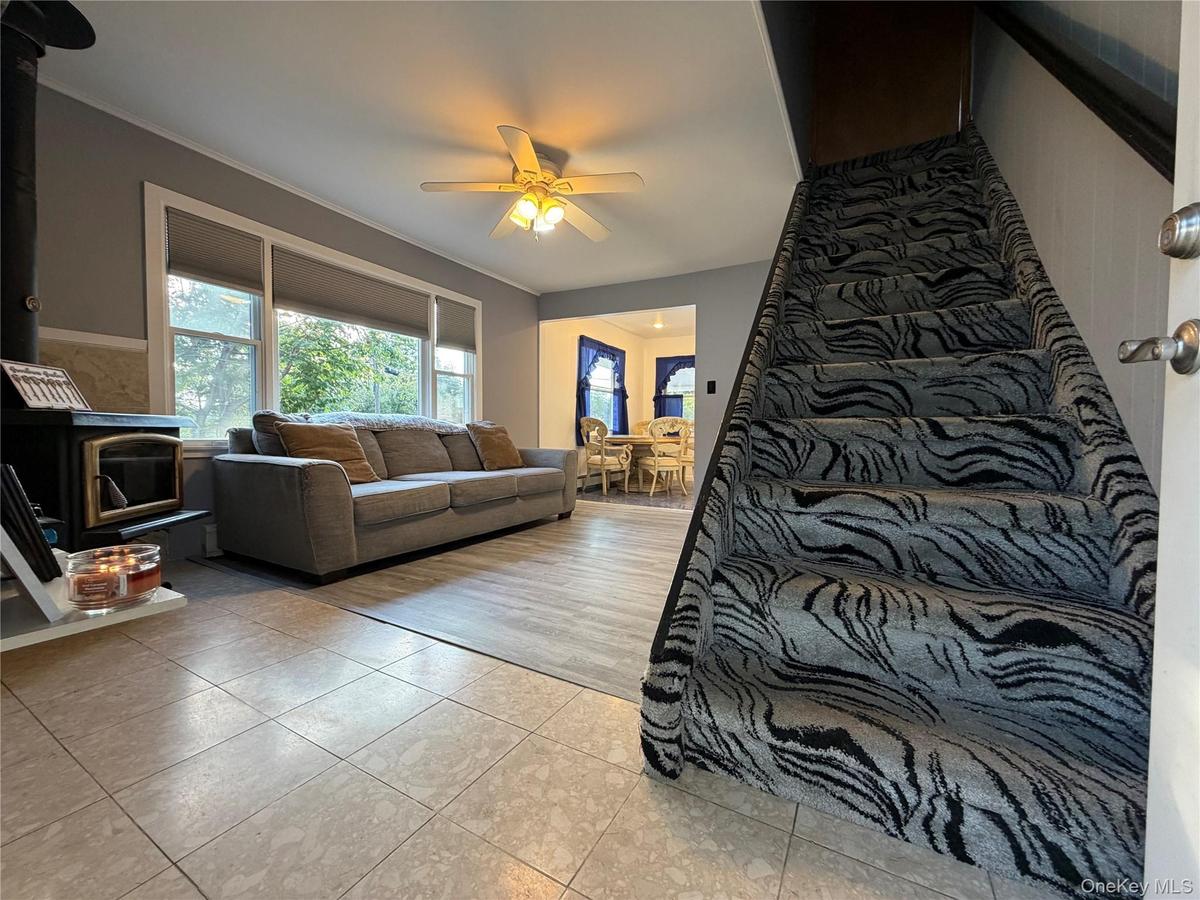 Wood-burning stove, large windows, ceiling fan, carpeted staircase, and tiled flooring