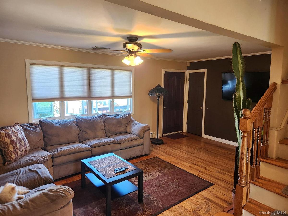 Hardwood flooring throughout, large window with cellular shades, ceiling fan with integrated lighting, dark wood-finish doors, and a wood staircase