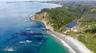 Aerial perspective of coastal landscape featuring sandy shoreline, rocky cliffs, dense forest canopy, and a natural inland pond