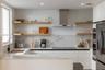 Modern kitchen featuring white flat-panel cabinetry, light-toned wood floating shelves, stone-surface countertops, full-height stone backsplash, and a