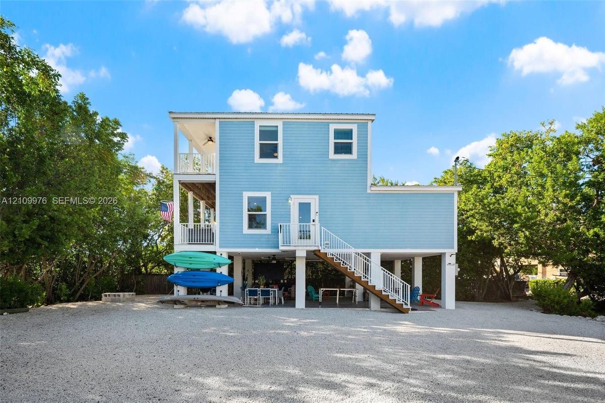 Elevated two-story residence featuring light blue lap siding, stacked covered porches, white metal railings, and a gravel ground-level parking area
