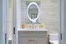 Custom vanity with stone-surface countertop, integrated rectangular sink, oval backlit mirror, and floor-to-ceiling stone-tiled wall
