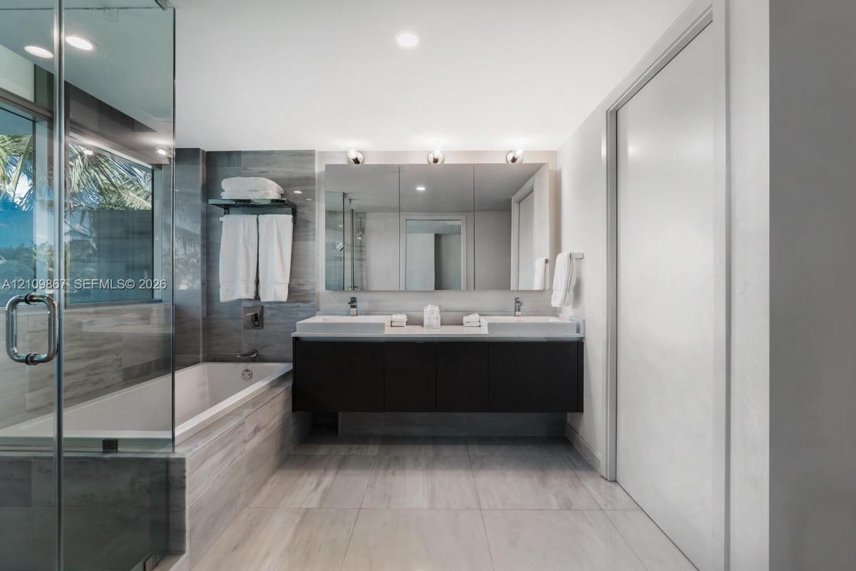 Floating dual-sink vanity with a countertop, expansive wall-mounted mirror, glass-enclosed soaking tub, and tile flooring