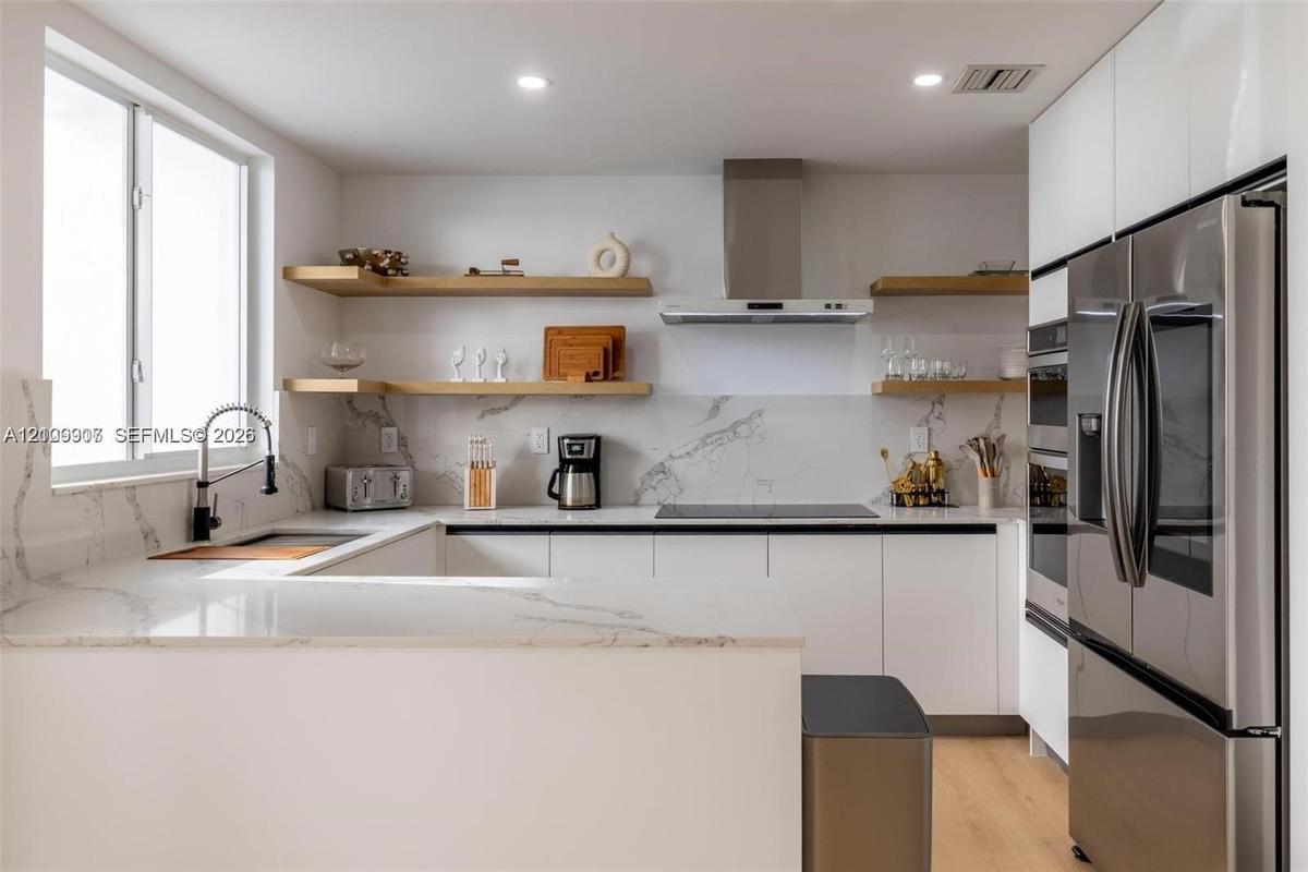 Modern kitchen featuring white flat-panel cabinetry, stone-finish countertops with matching backsplash, built-in stainless steel appliances, open wood
