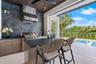 Covered outdoor kitchen featuring a dark stone backsplash, stainless steel range hood, integrated gas grill, flat-panel cabinetry, and dark-toned ston