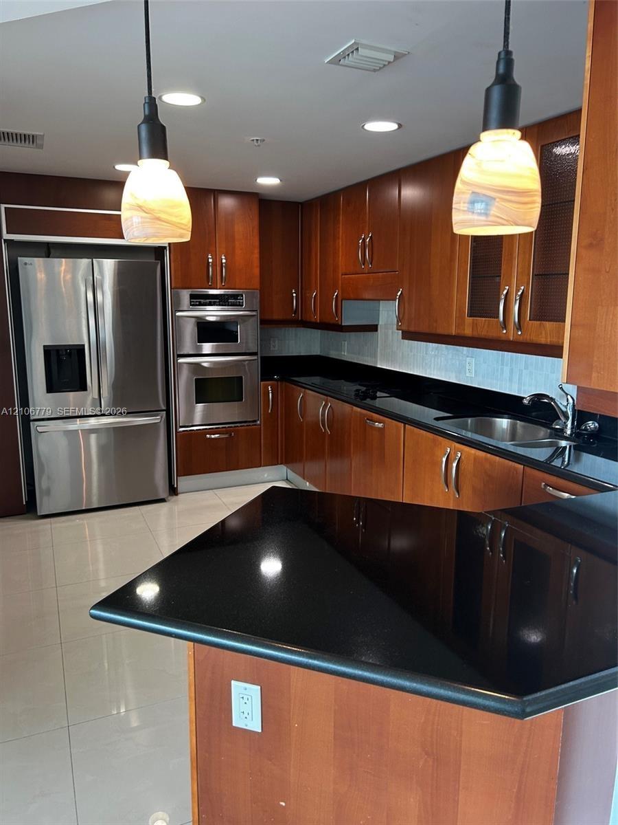 Kitchen featuring wood cabinetry, black countertops, stainless steel built-in double oven, stainless steel refrigerator, and pendant lighting