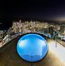 Rooftop infinity-edge pool featuring integrated lighting, tiled decking, and stainless steel handrails, overlooking an illuminated urban skyline