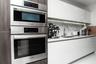 Modern kitchen featuring a stacked stainless steel Bosch wall oven and microwave, high-gloss white cabinetry, stone-finish backsplash, and an undermou
