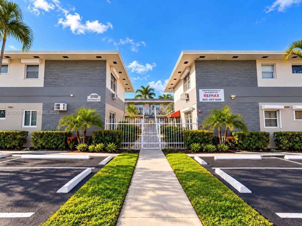 Multi-unit exterior featuring gray stone-veneer accents, paved parking area, gated pedestrian entrance, and manicured landscaping